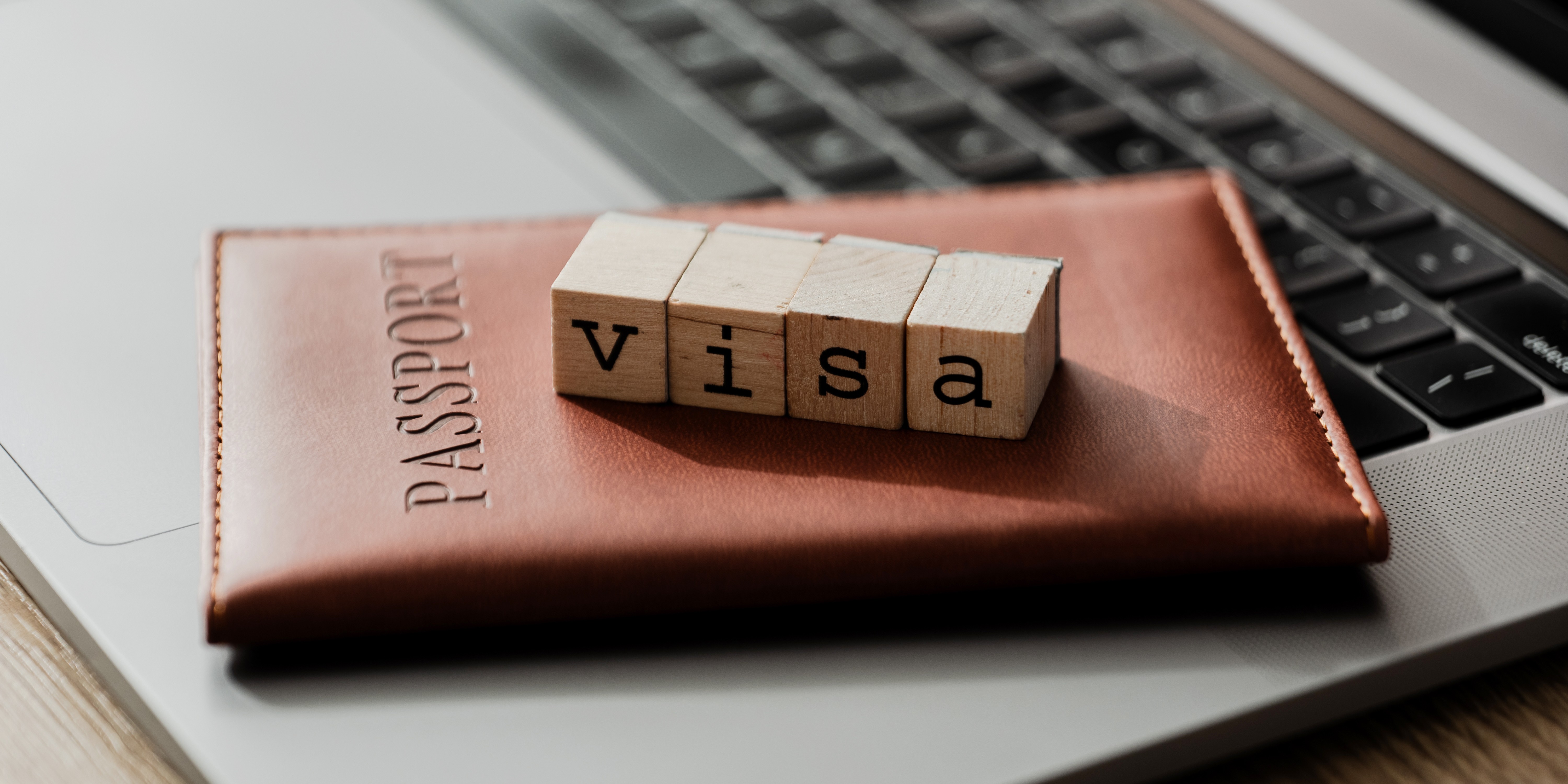 A passport on top of a laptop, with wooden blocks that spell "visa" stacked on top of the passport.